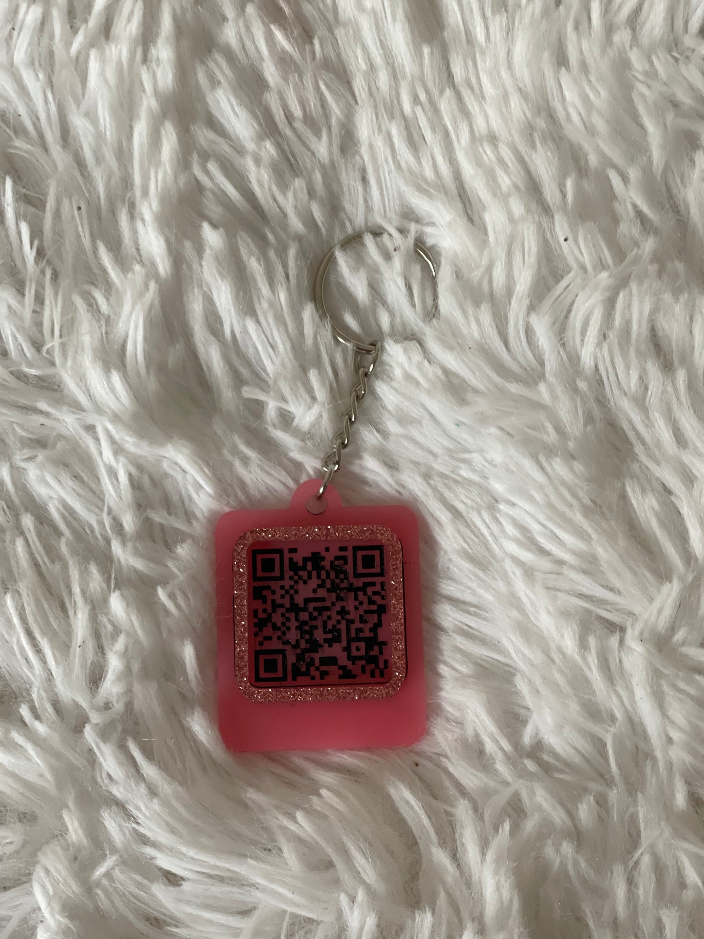 A pink QR code keychain with glitter, placed on a white furry surface.