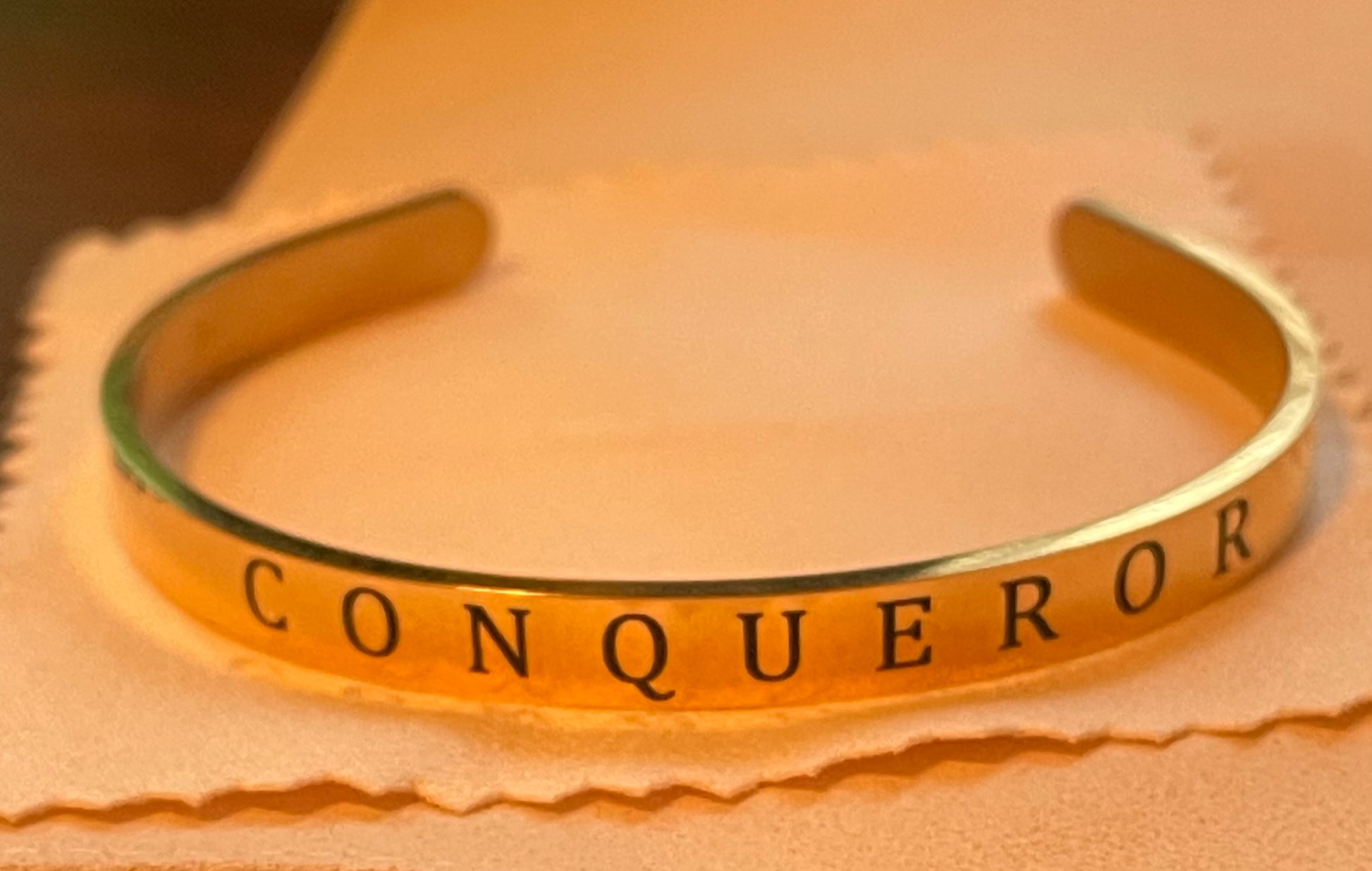 Gold 1/4 inch  bracelet with 'CONQUEROR' engraving on a beige background