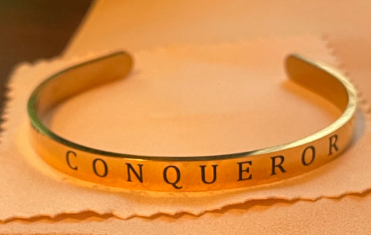 Gold 1/4 inch  bracelet with 'CONQUEROR' engraving on a beige background