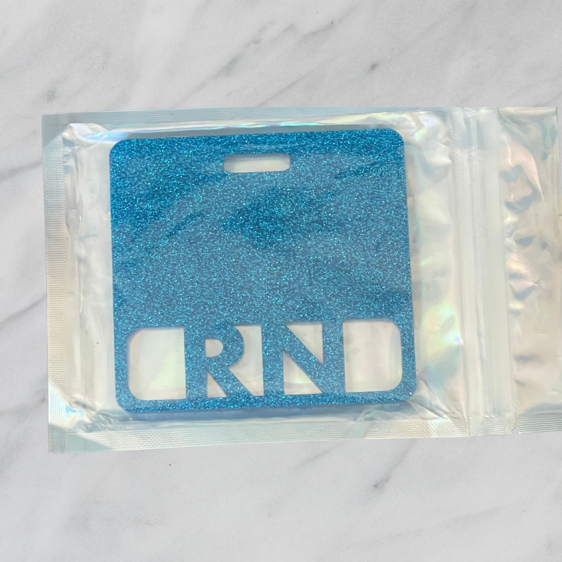 Blue glittery 'RN' badge mate in clear packaging on a marble surface