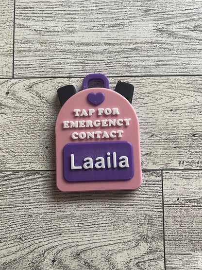Pink backpack-shaped emergency contact tag with name on a wooden surface