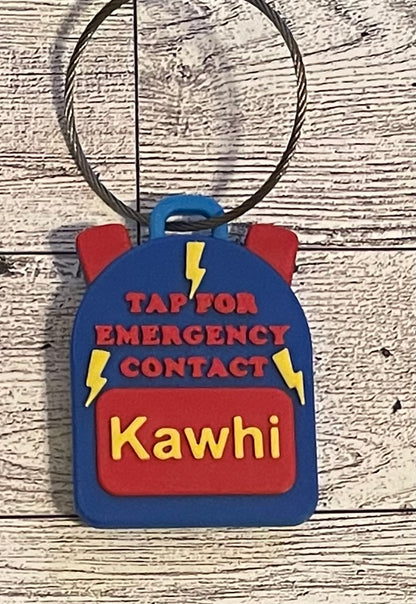 Backpack-shaped keychain with 'Tap for Emergency Contact' text on a wooden surface