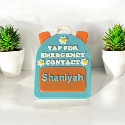 Backpack-shaped emergency contact device with 'Tap for Emergency Contact name text, surrounded by potted plants.