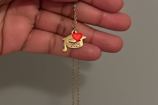 A stainless steel graduation cap pendant necklace with a red heart and the year 2025 inscribed on the pendant. The necklace is held between fingers, showing its length.