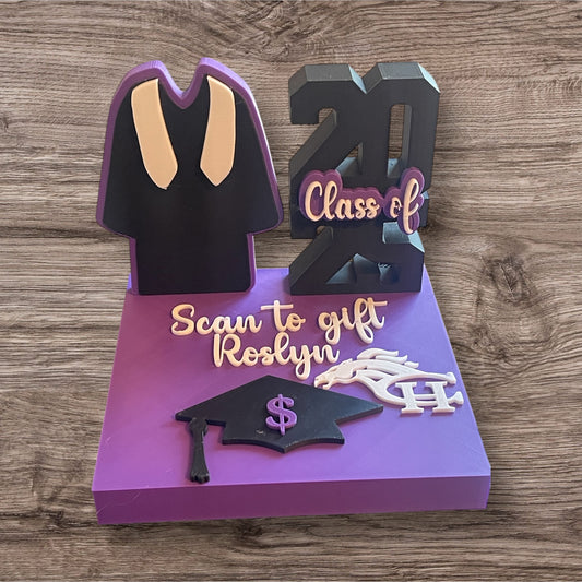 A 3D printed display stand with graduation-themed elements, including a graduation cap and gown, and personalized text areas for a name and school mascot or initials. The stand is in purple and black colors with a 'Scan to gift' text feature.