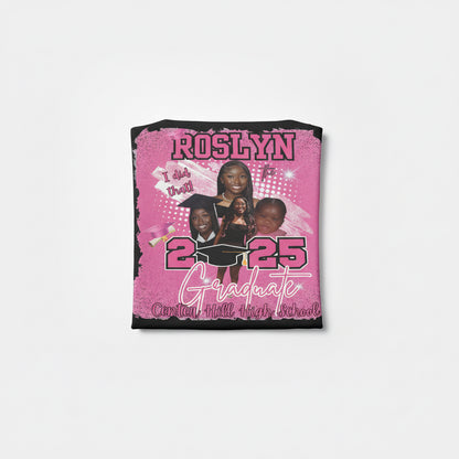 Pink graduation-themed shirt with text and images of people, held against a brick wall.