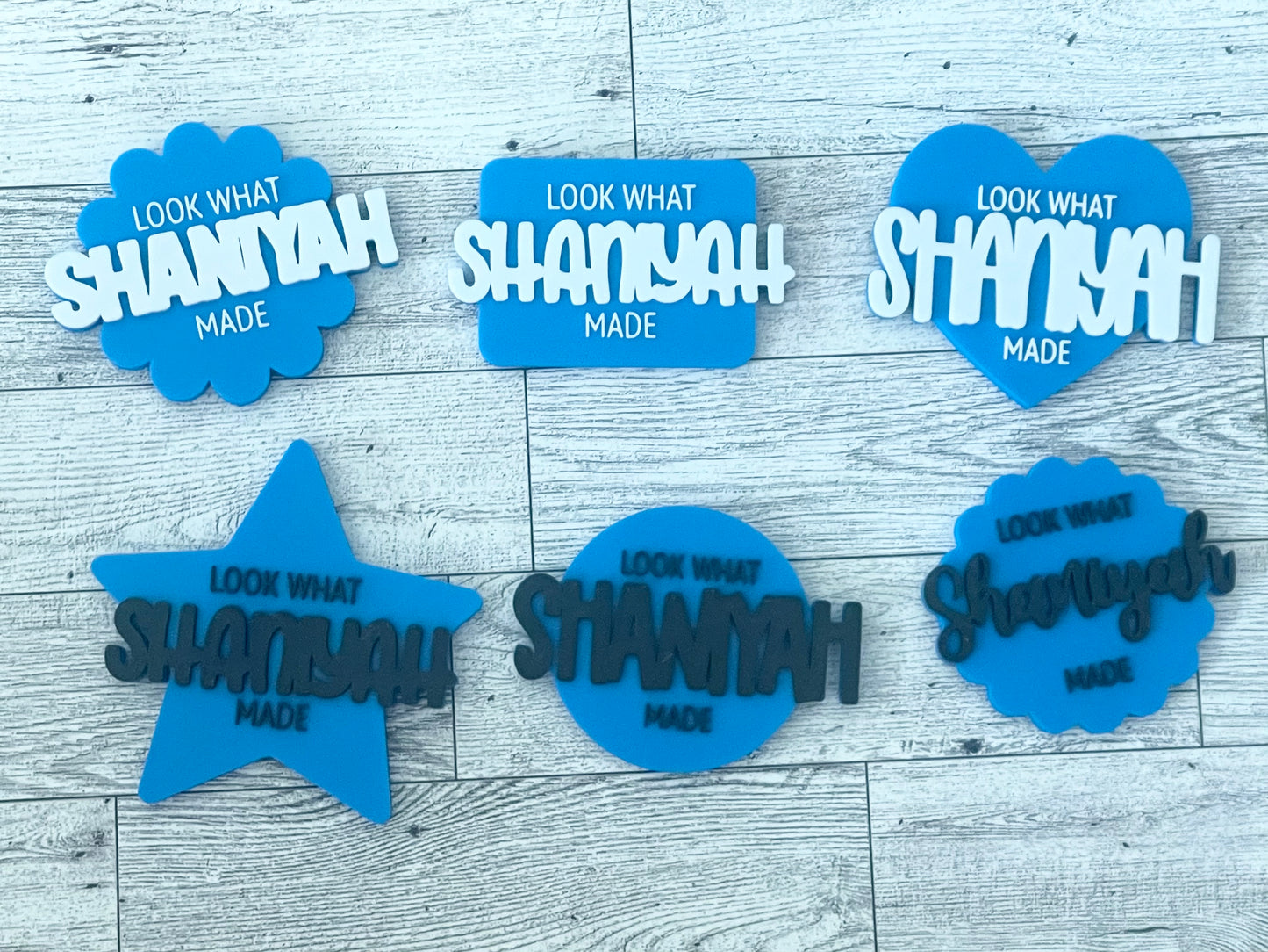 Set of blue and white decorative signs with 'Look what Shaniyah made' text on a wooden surface.