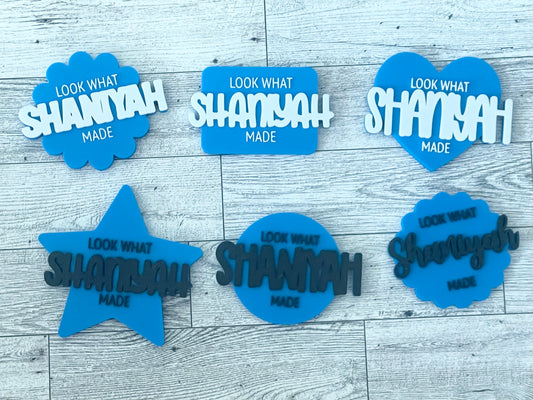 Set of blue and white decorative signs with 'Look what Shaniyah made' text on a wooden surface.