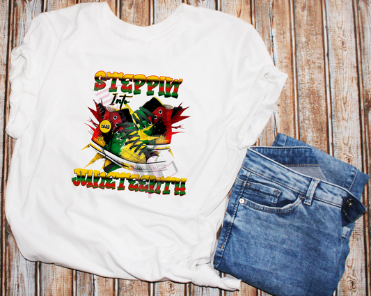 A white t-shirt with a graphic print featuring the text 'STEPPIN' INTO FREEDOM - JUNETEENTH' and images of sneakers, denoting a theme related to the Juneteenth holiday.