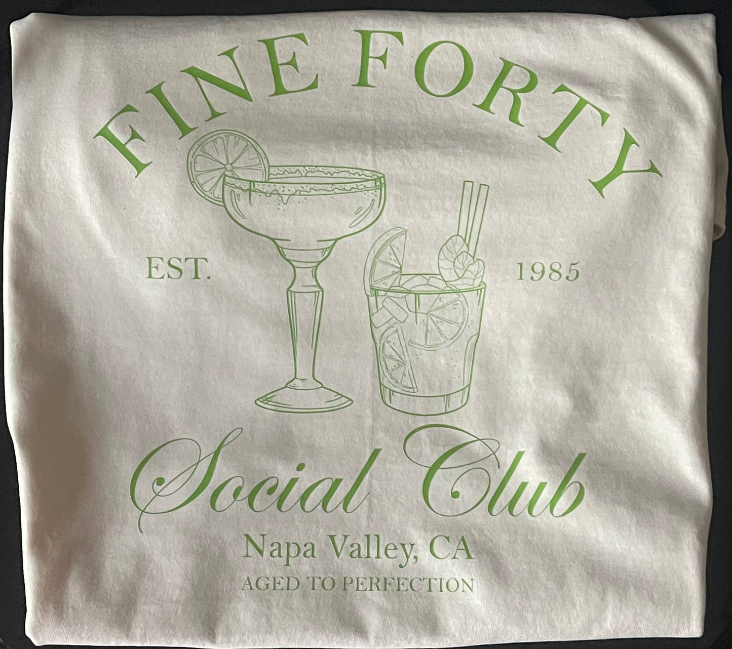 Folded white shirt with green text and cocktail illustrations on a black background