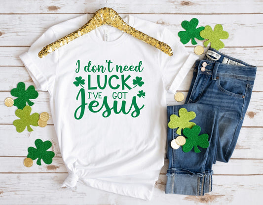A white t-shirt with the text 'I DON'T NEED LUCK I'VE GOT JESUS' printed in green, accompanied by shamrock decorations and placed on a wooden surface with a pair of blue jeans and a gold-colored hanger in the background.