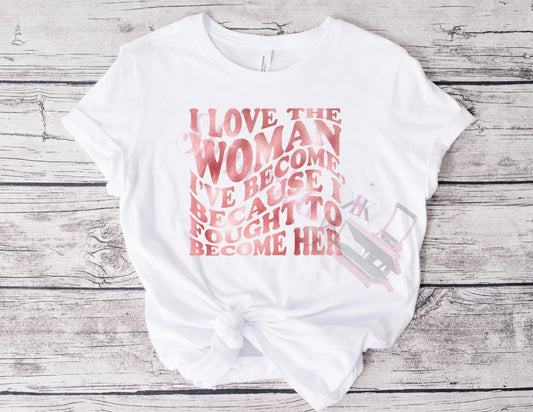 A white t-shirt with a rose gold text graphic reading 'I love the woman I'm becoming because I fought to become her'
