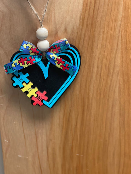 A wooden car charm painted with a colorful autism-themed design, featuring a heart shape with puzzle pieces and a bow.