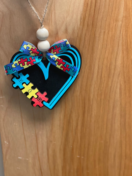 A wooden car charm painted with a colorful autism-themed design, featuring a heart shape with puzzle pieces and a bow.