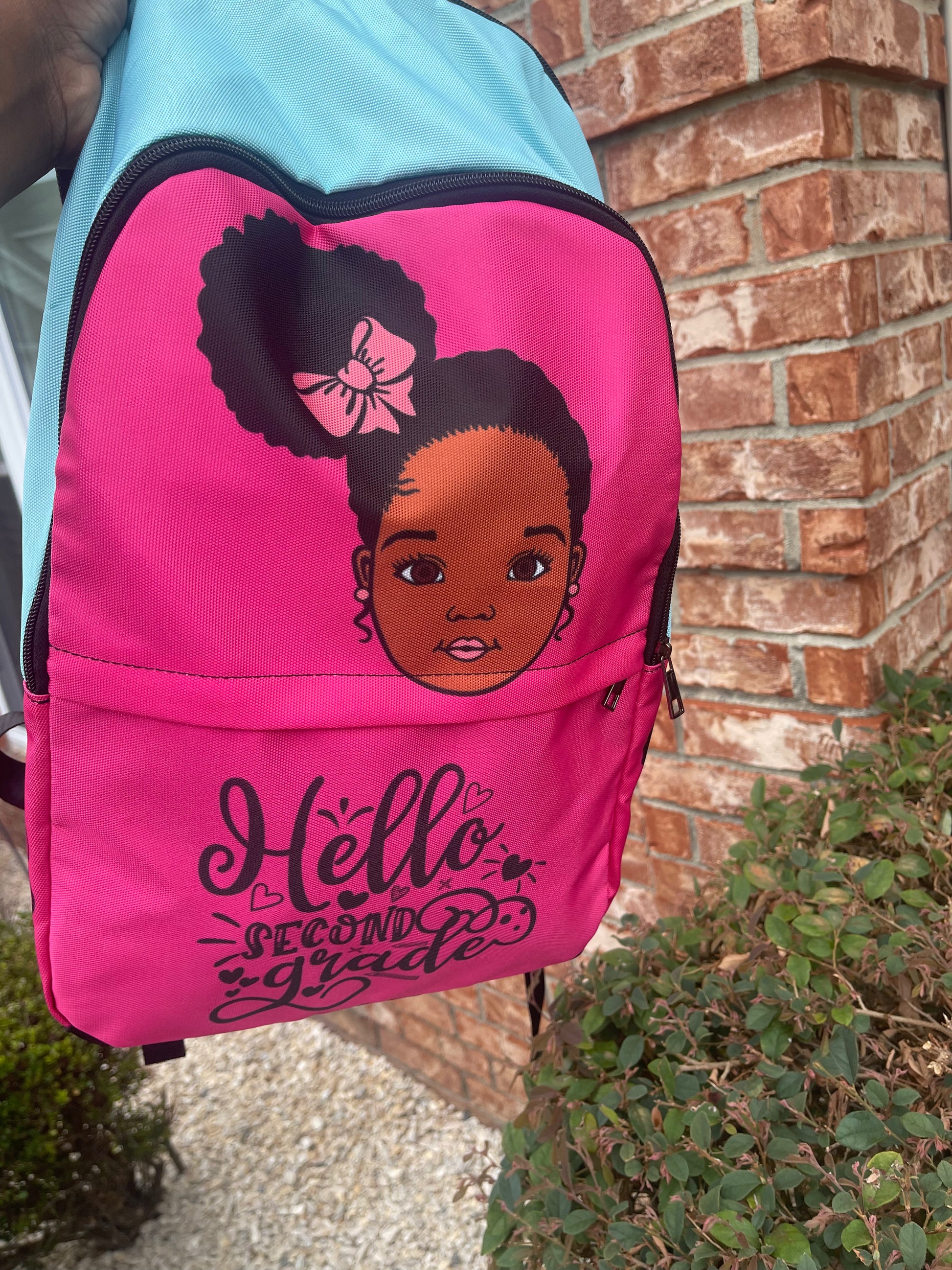 A pink custom backpack featuring a black and white illustration of a character's head on the front, with the text 'Hello, Second Grade' and a bow design.