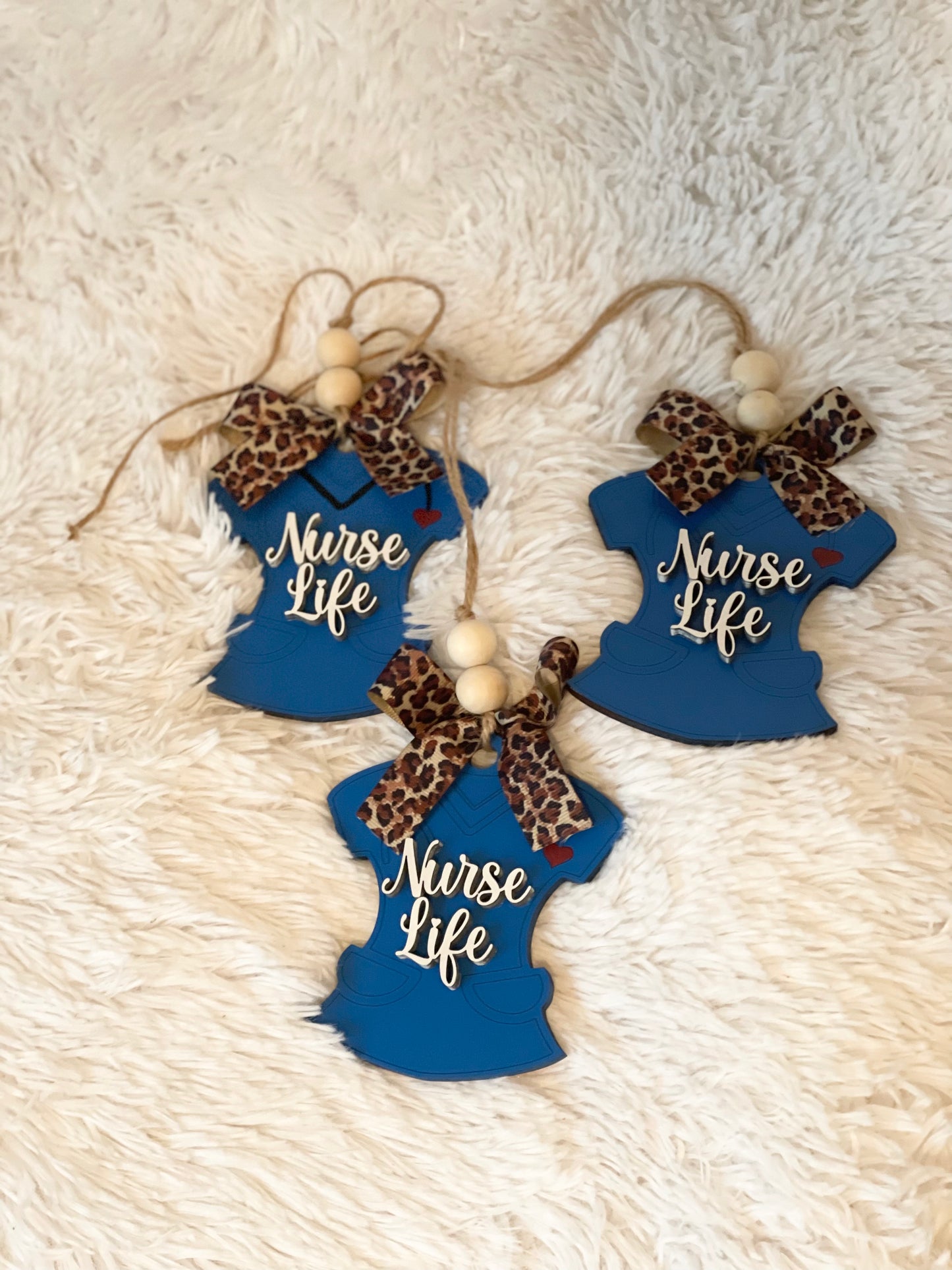Two blue wooden car charms with 'Nurse Life' text and a leopard print bow, displayed on a white furry surface.